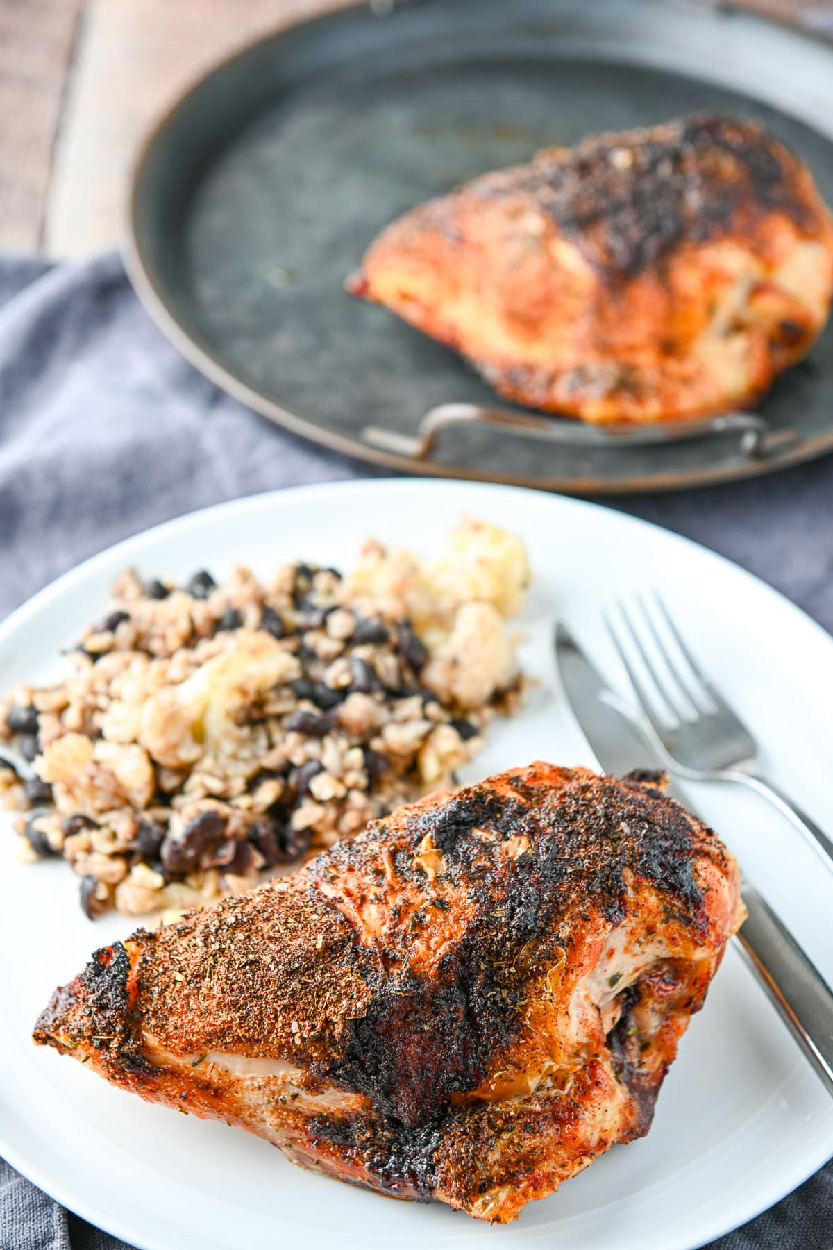 A white plate with the cooked chicken breast on it with rice and a metal tray in the back with another breast on it