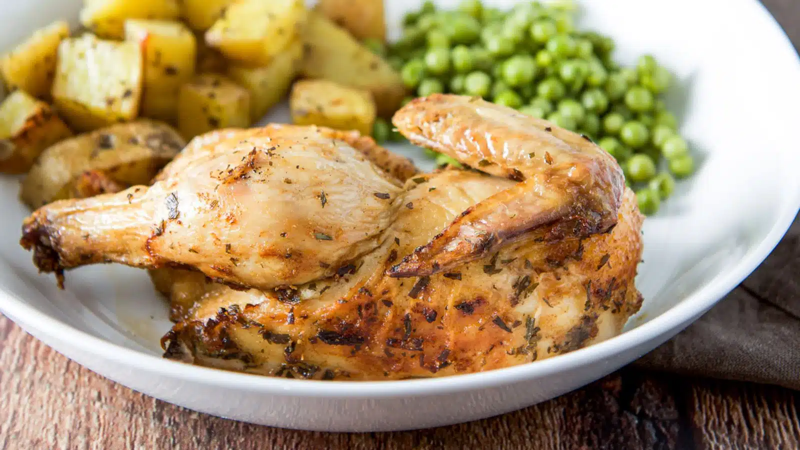 A white deep plate with half a hen on it with potatoes and peas