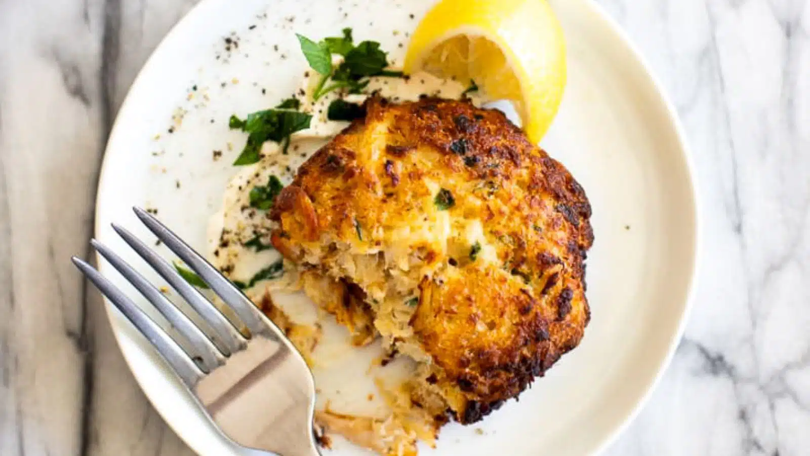 a white plate with a crab cake, lemon, and a fork