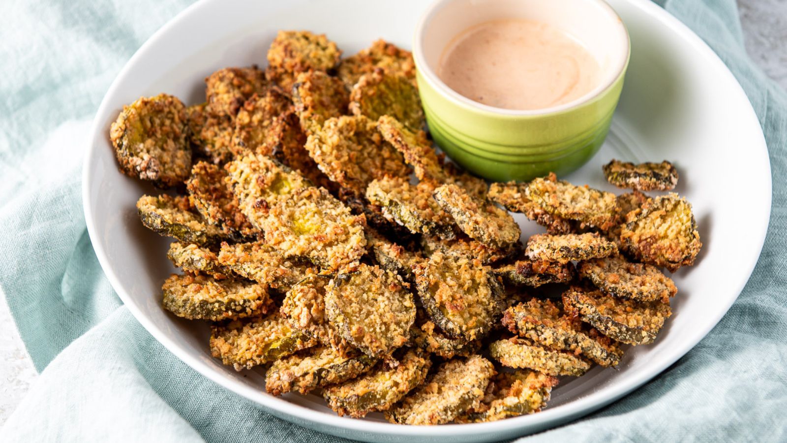A deep white plate with fried pickles on it along with a sauce in a small cup