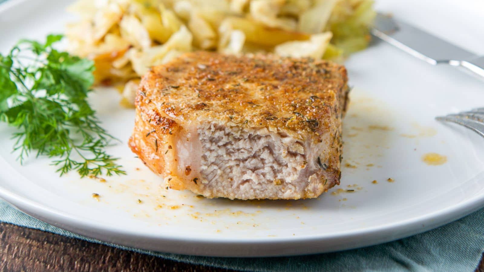 A white plate with a piece cut out of a pork chop with herbs and cabbage in the back