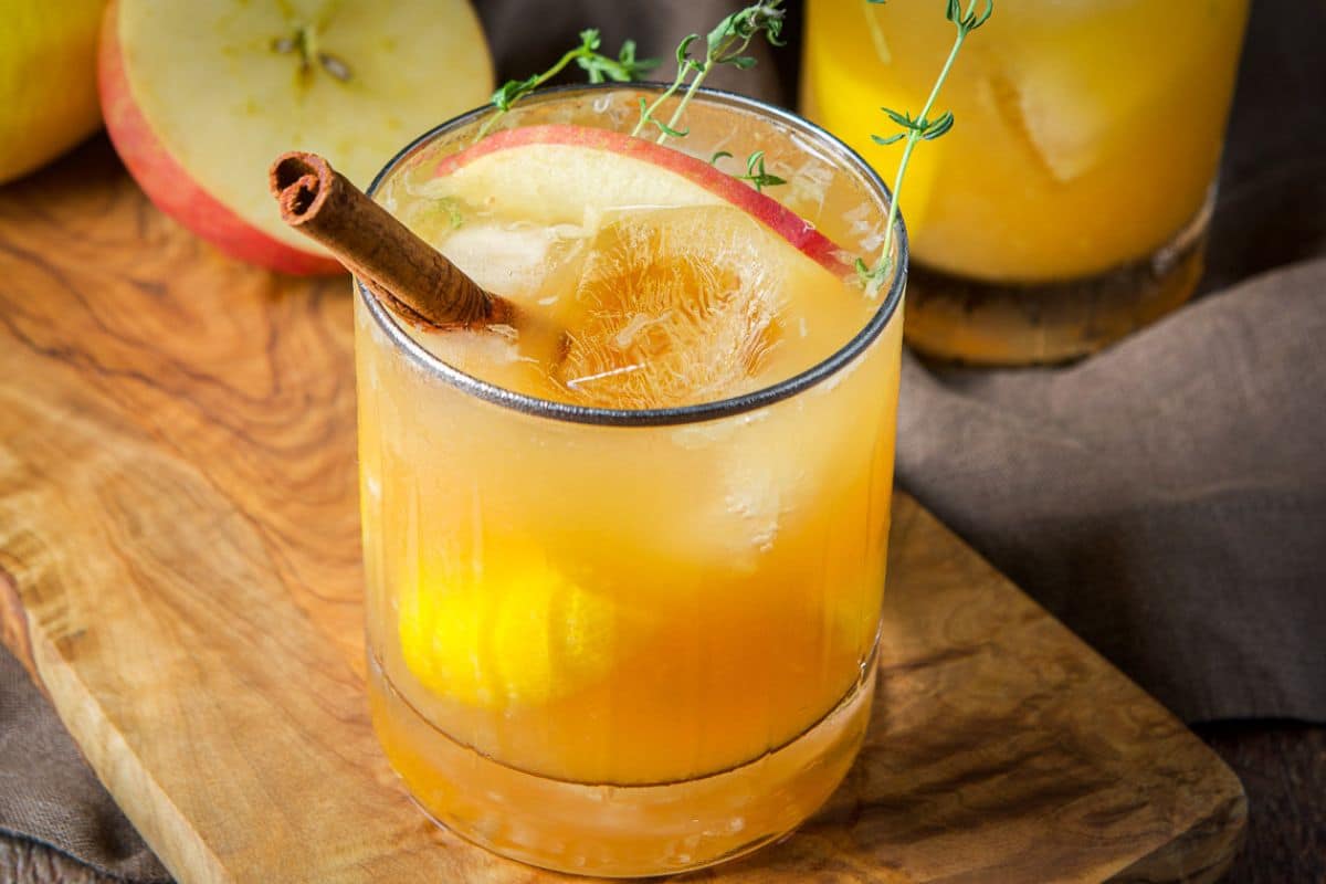 A glass on a board filled with the apple drink with a sliver of apple, cinnamon stick, and herbs