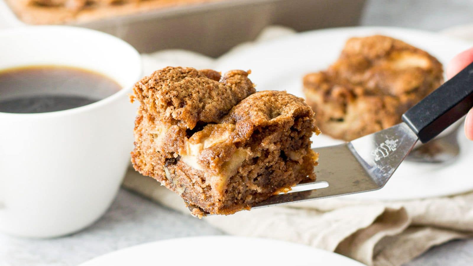 A spatula holding up a piece of apple cake with coffee to the side and another piece in the back