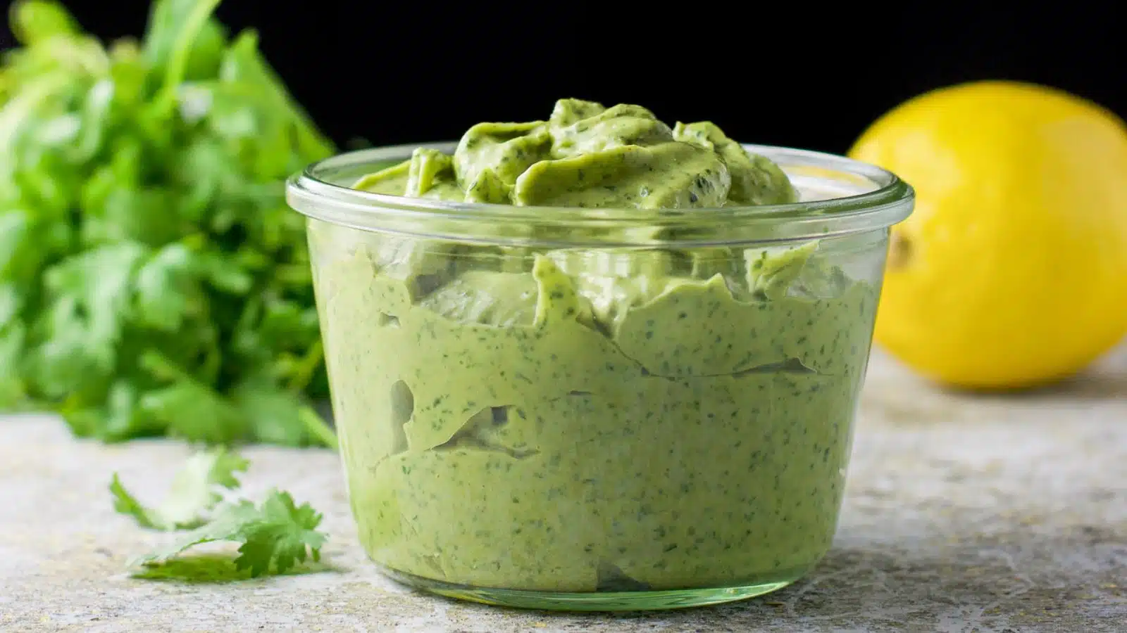 A wide mouthed glass jar with a green dip in it with cilantro and a lemon in the back