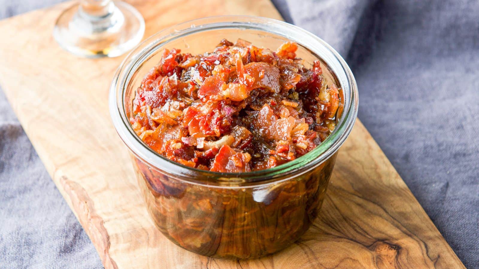 A wide glass jar with the bacon mixture in it on a wooden board