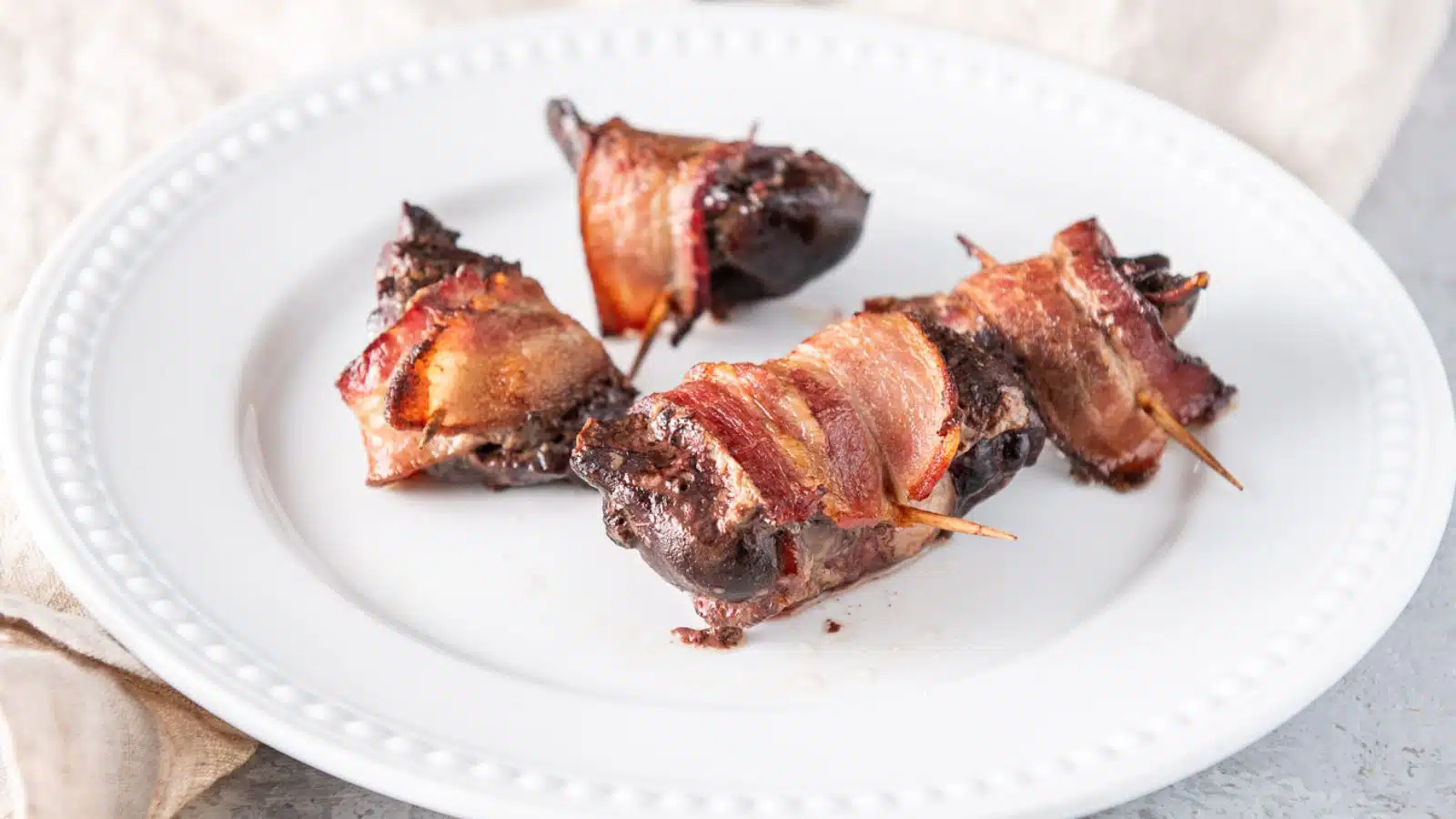 a white beaded plate with four chicken livers wrapped with bacon and toothpicks through them