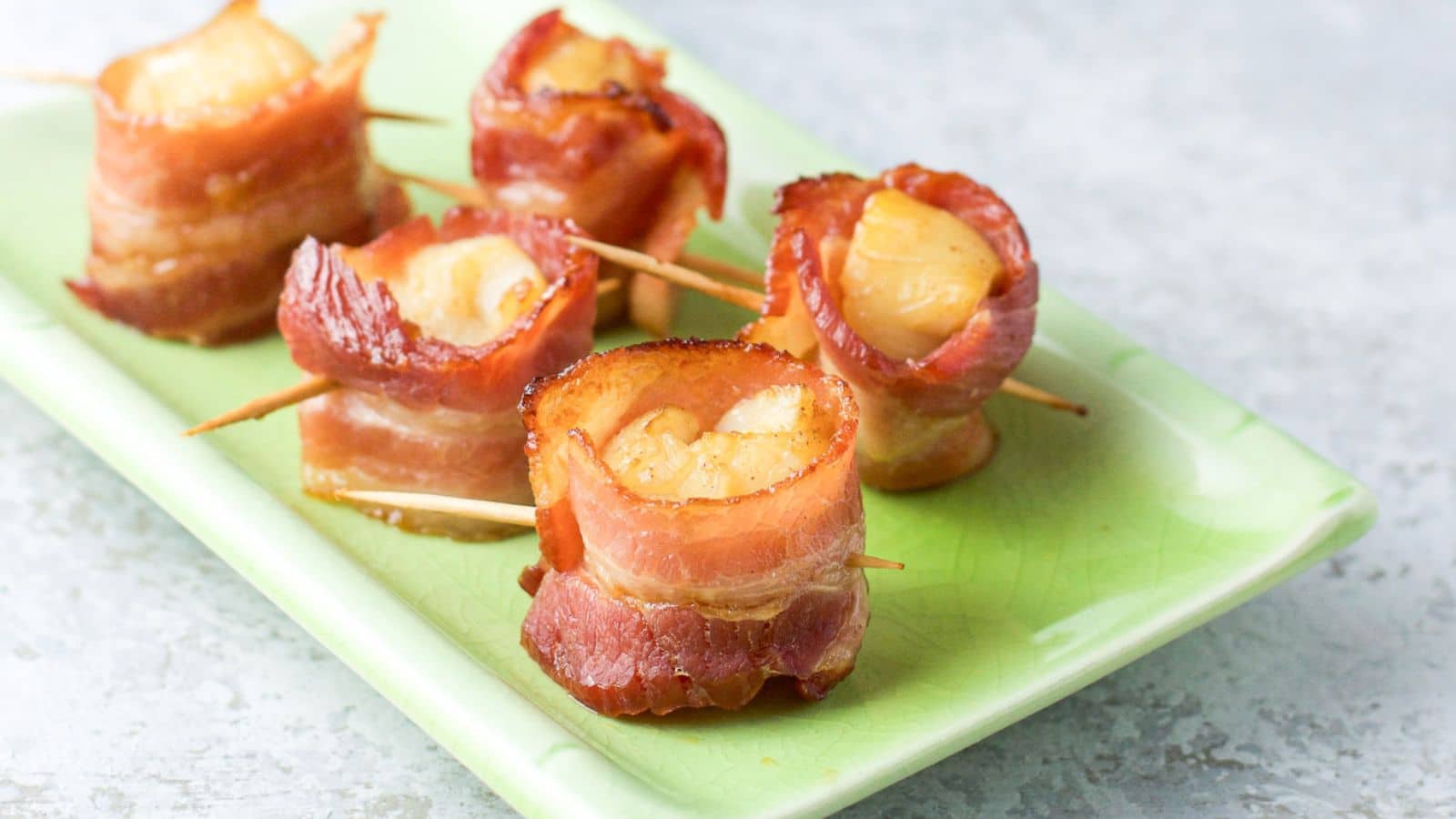 A little green rectangular plate with five bacon wrapped scallops with toothpicks out of it