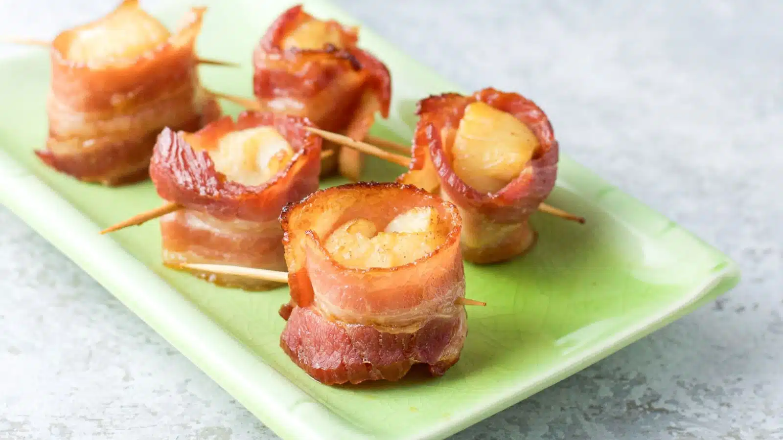 A little green rectangular plate with five bacon wrapped scallops with toothpicks out of it