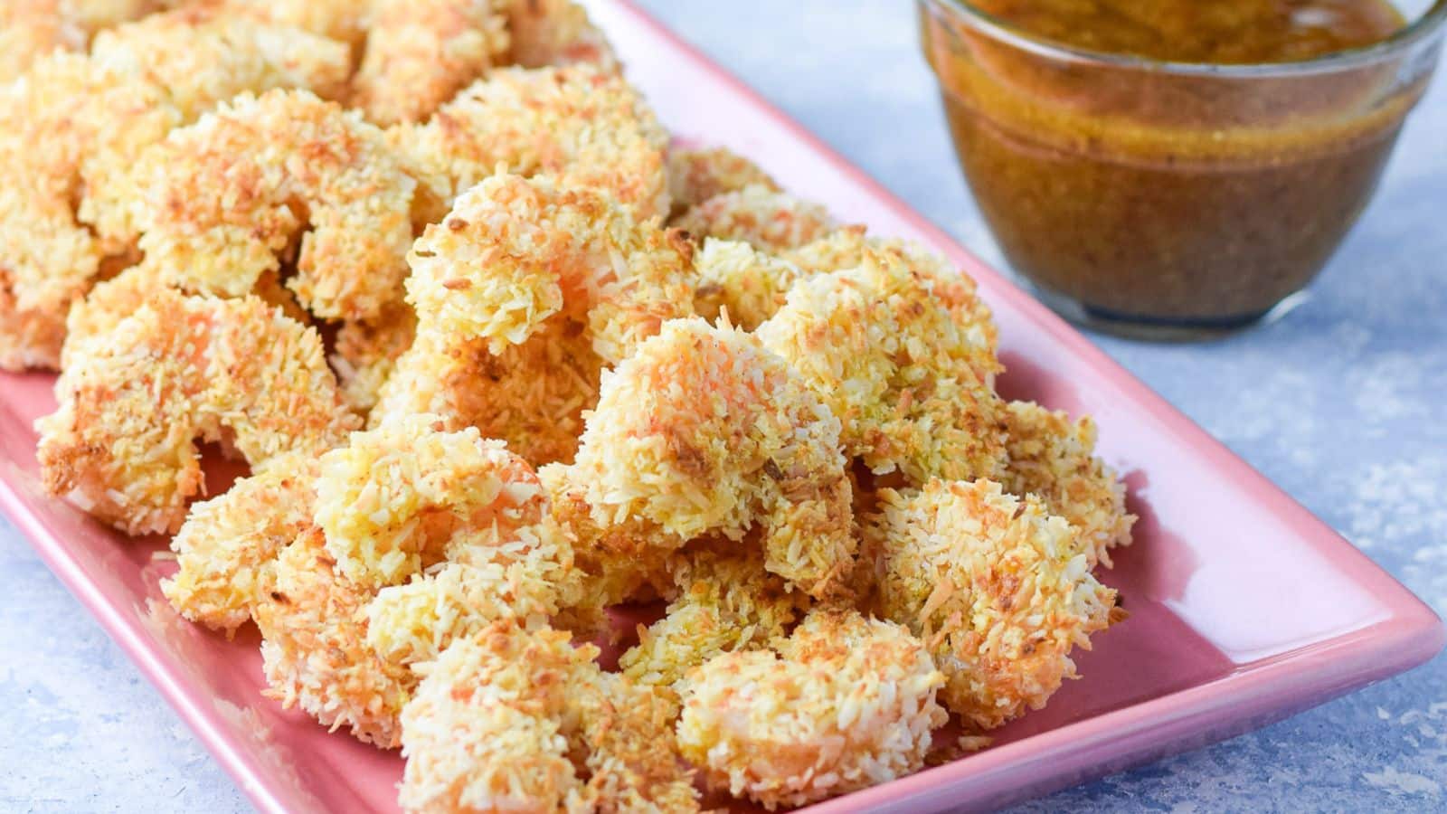 A pink platter with coconut shrimp on a blue board with dip next to it