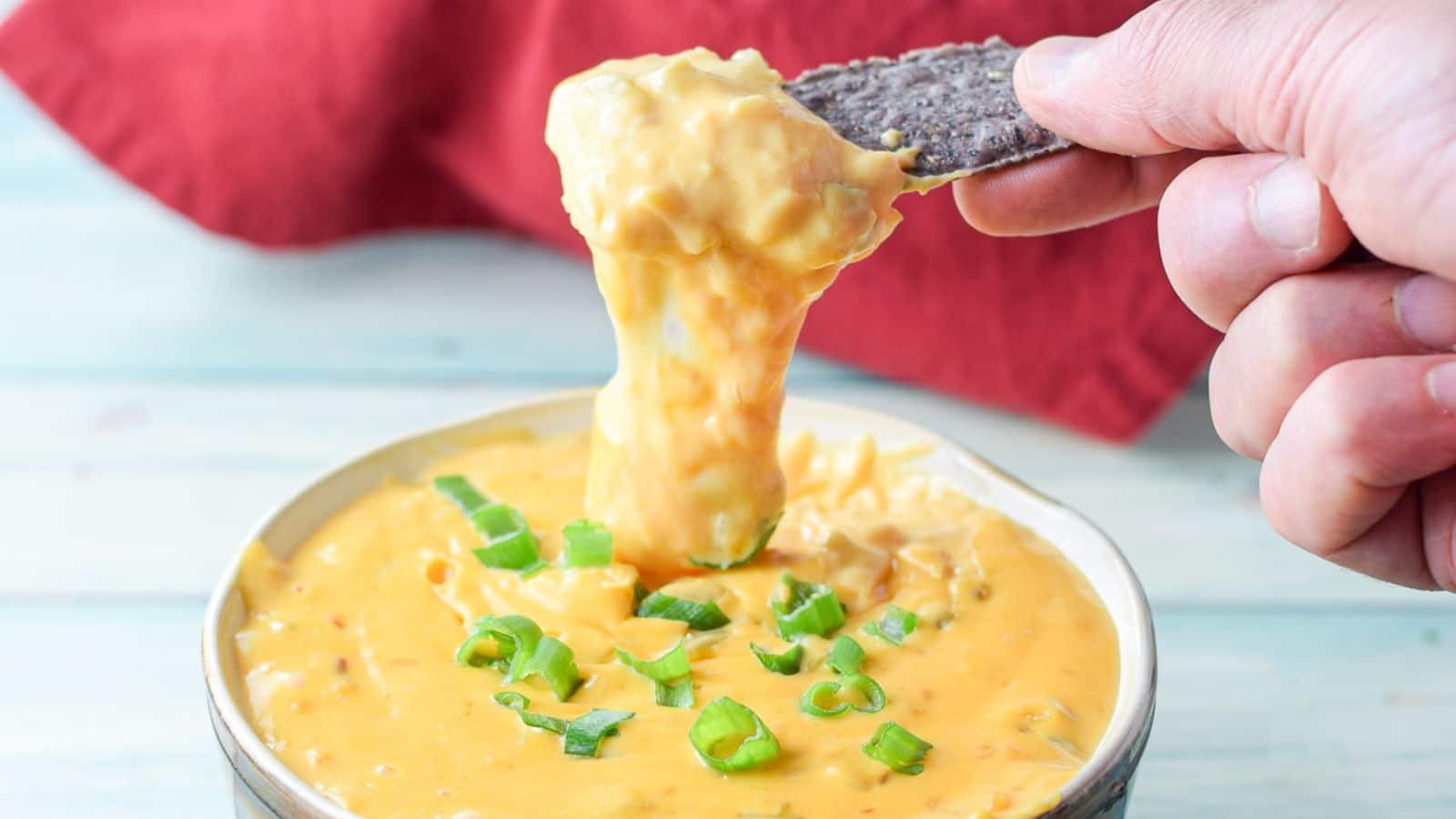 a hand holding a chip above a cheesy dip with the stings of cheese dropping down