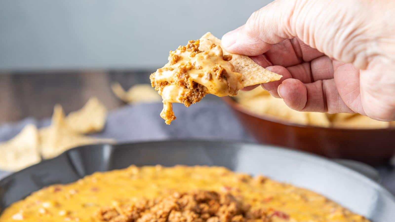 A hand holding a triangle tortilla chip filled with a cheese dip with chorizo