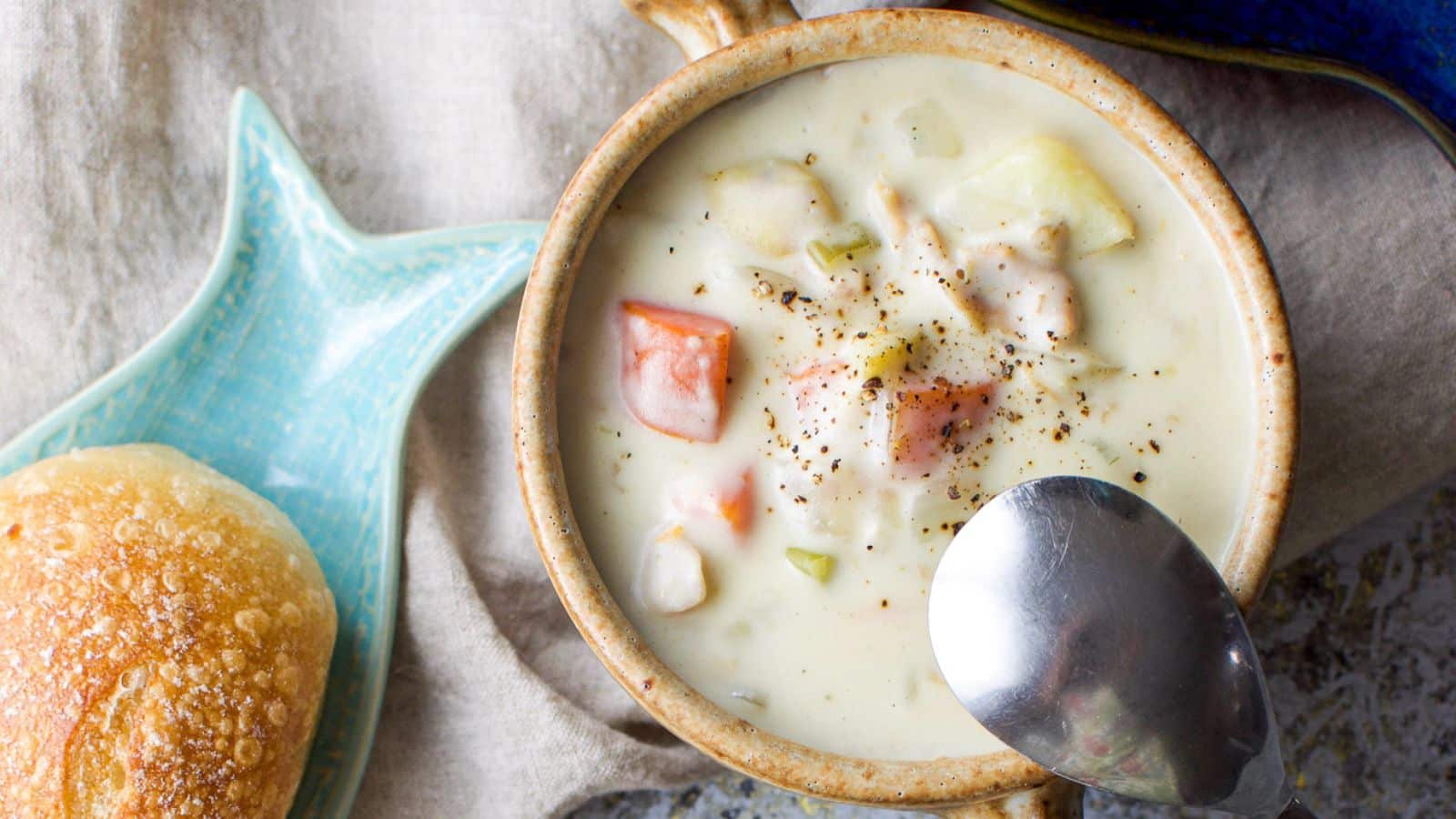 a bowl of chowder with black pepper and a roll on the side