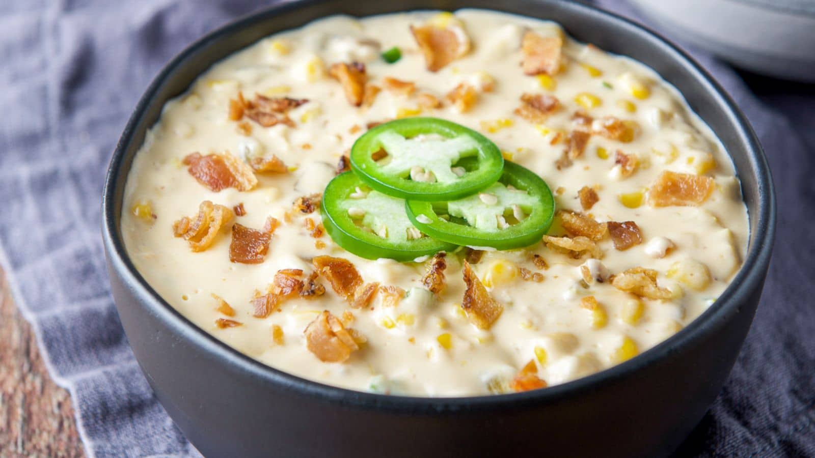A black bowl filled with a dip with corn, sprinkled on top is bacon and sliced jalapeno