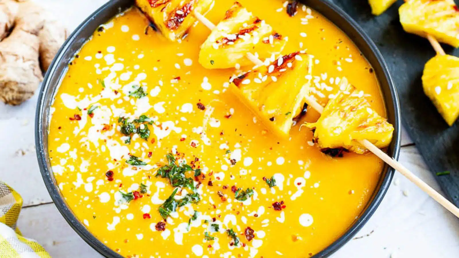 black bowl with a vibrant yellow-orange soup with grilled pineapple and herbs on top