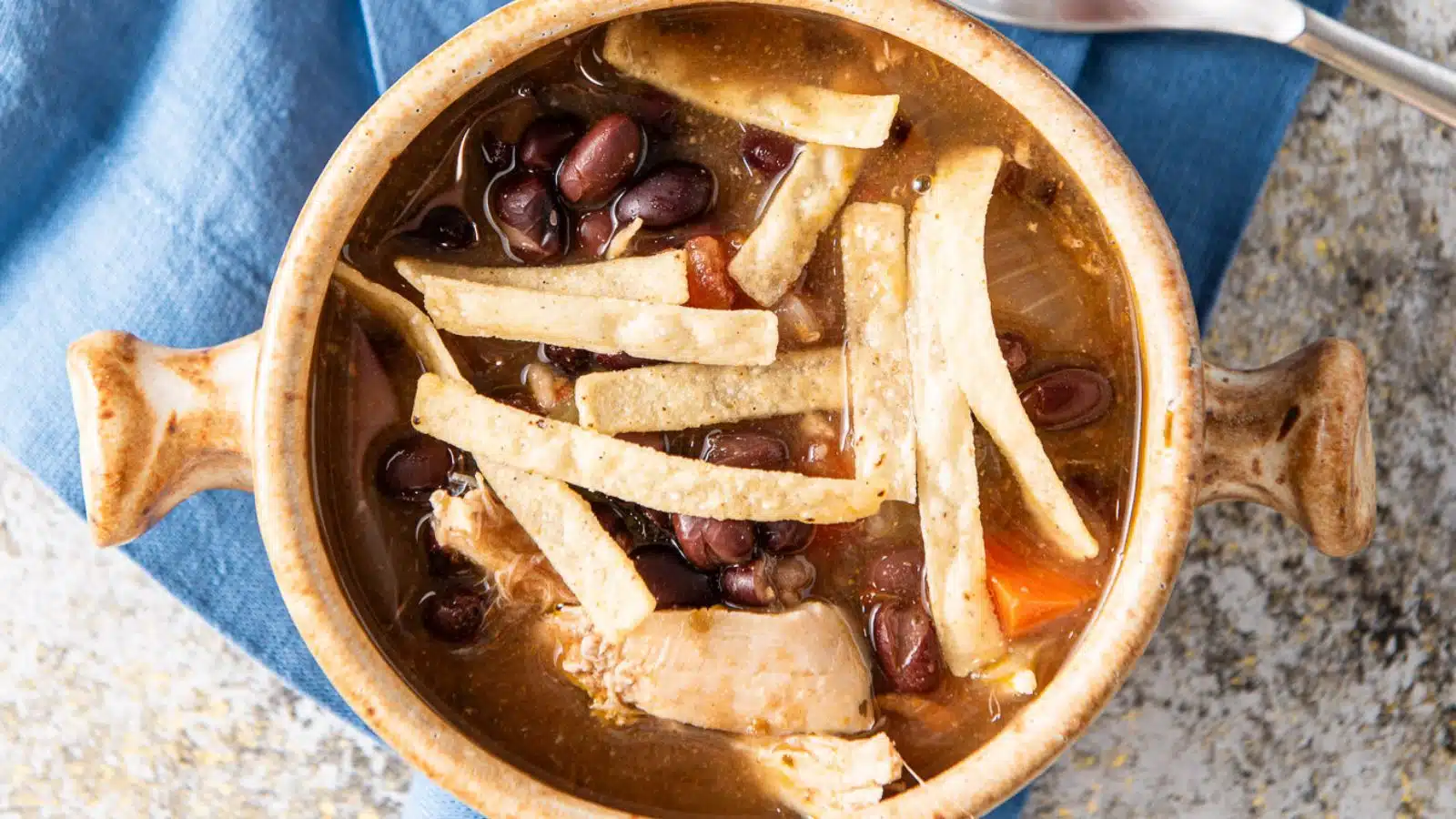 an overhead view of a two-handled soup bowl filled with the soup with beans, chicken and tortilla chip strips on top