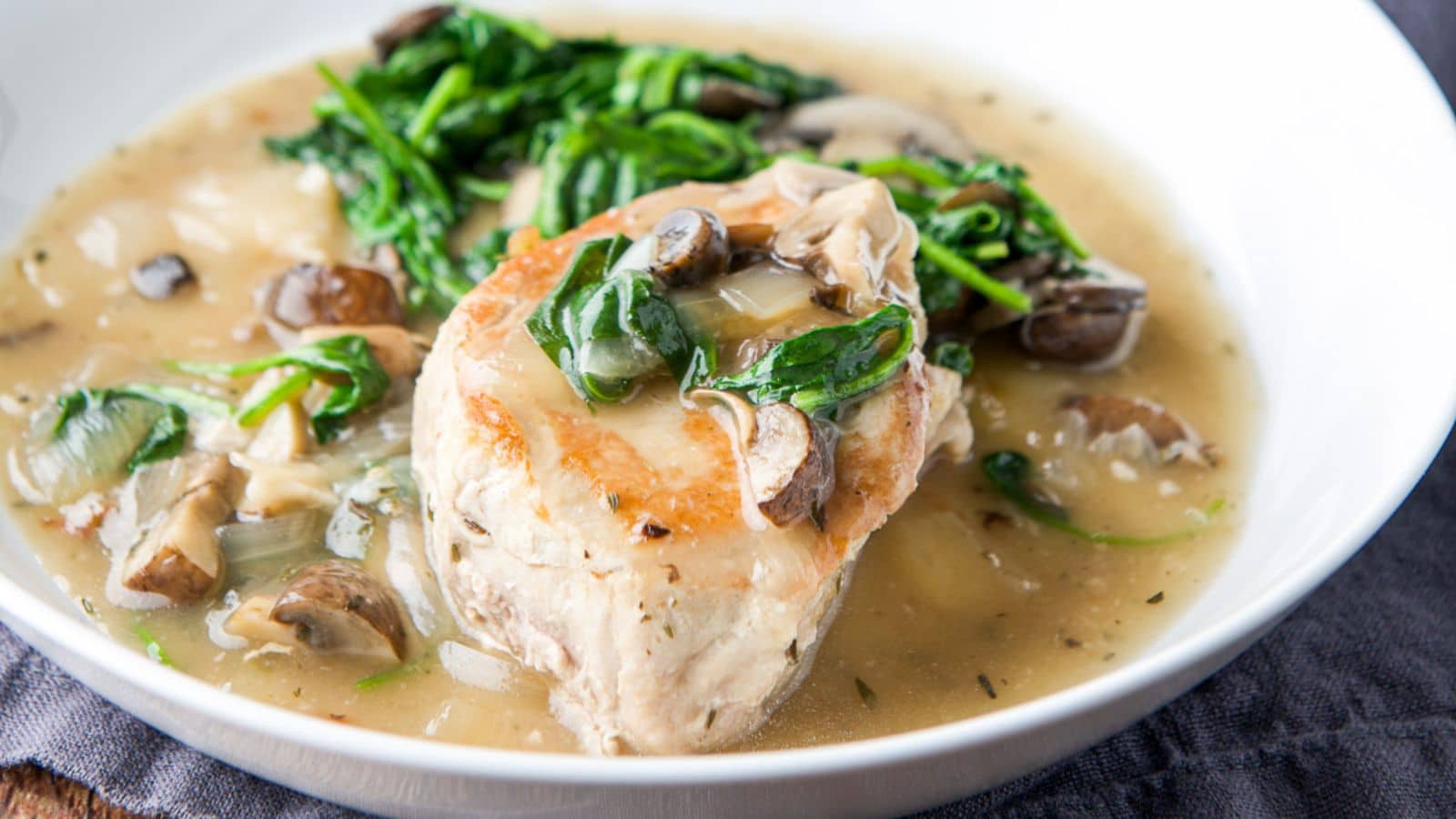 A white deep plate of a pork chop with brown gravy in it with vegetables