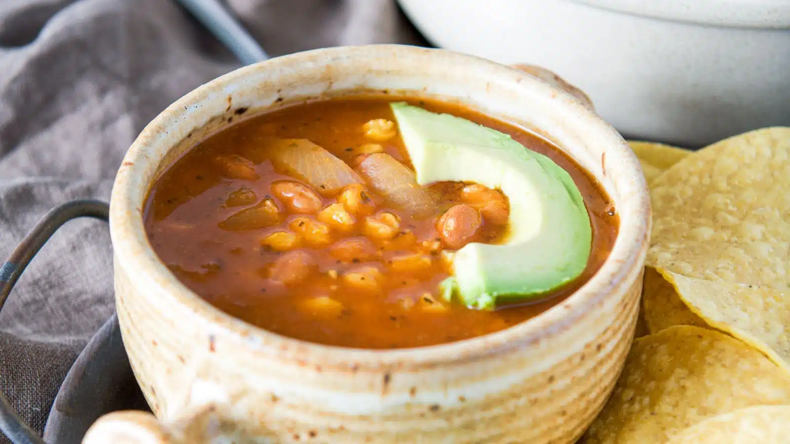 an avocado slice in on a tomato-based soup in a bowl with chips on the side