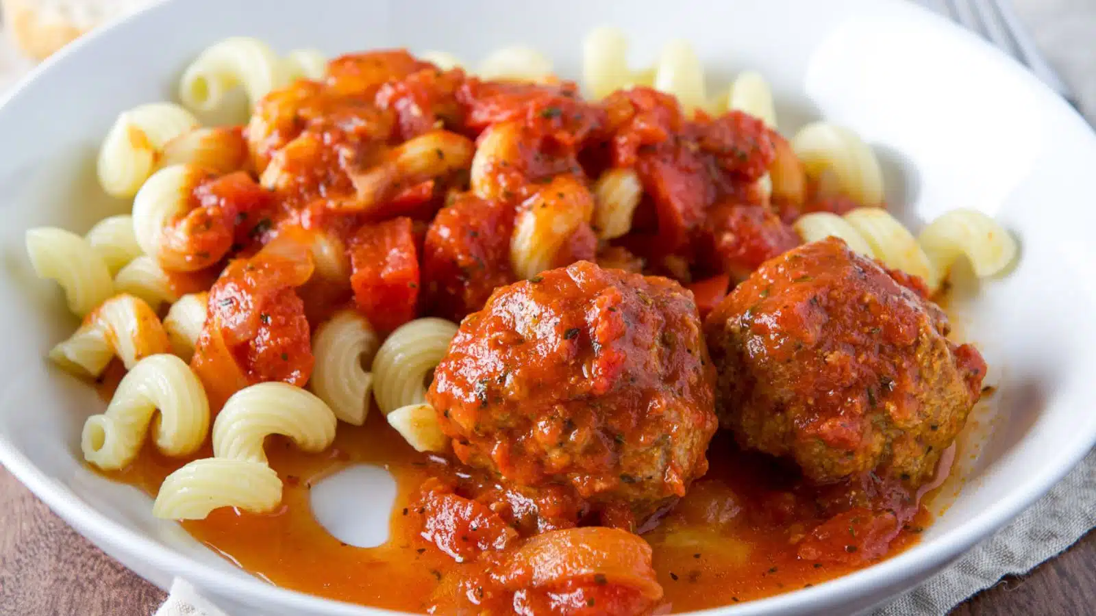 A white deep bowl with curly pasta with meatballs and gravy