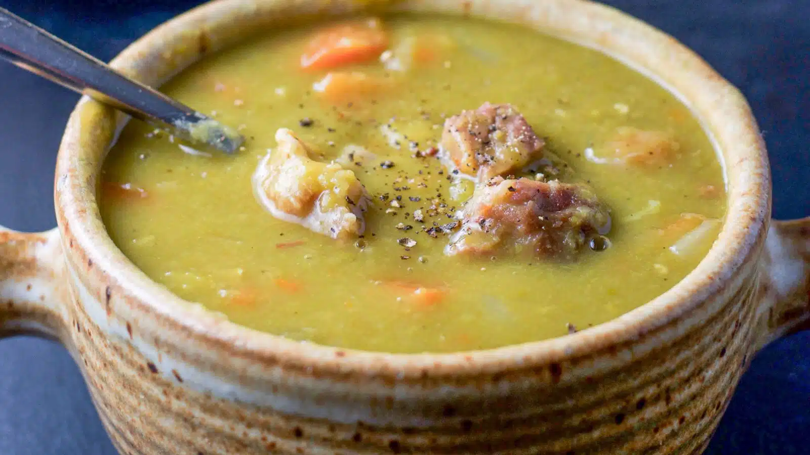 A crock with pea soup with ham in it, along with ground pepper and a spoon