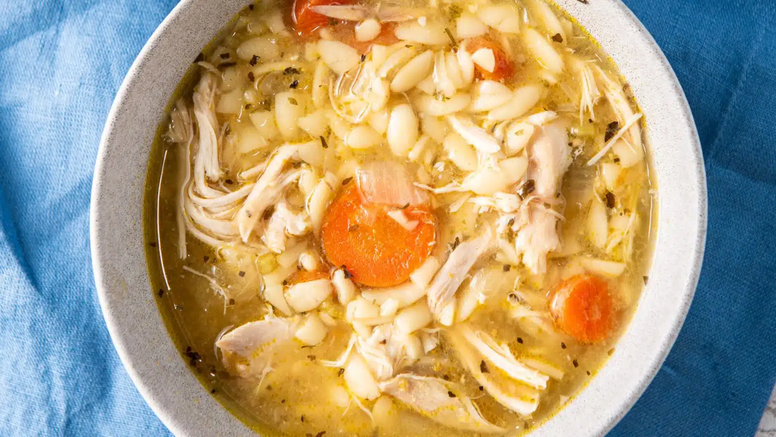 a close up of chicken soup with orzo and carrots in a grey bowl on a blue napkin