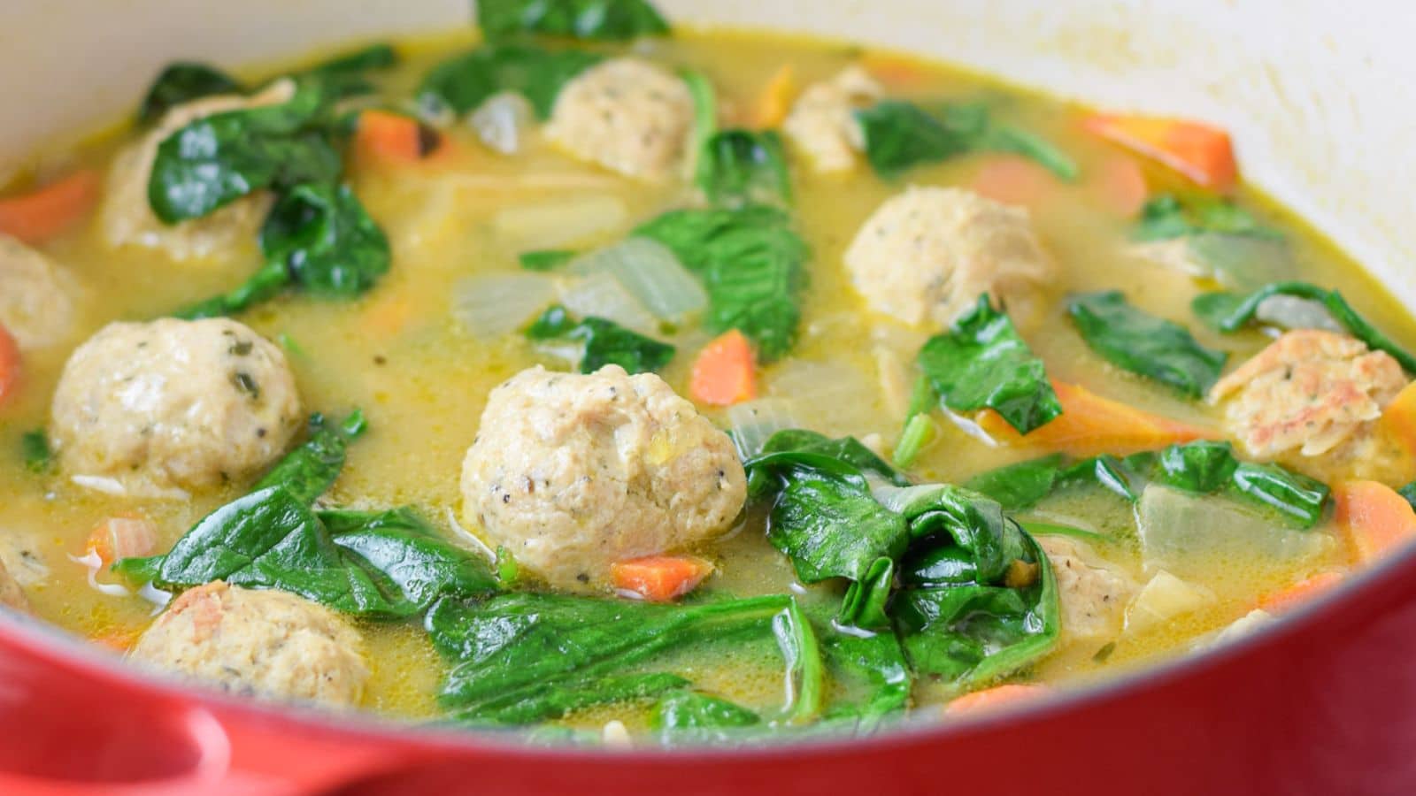 A red pan with a soup of meatballs, spinach, and vegetables