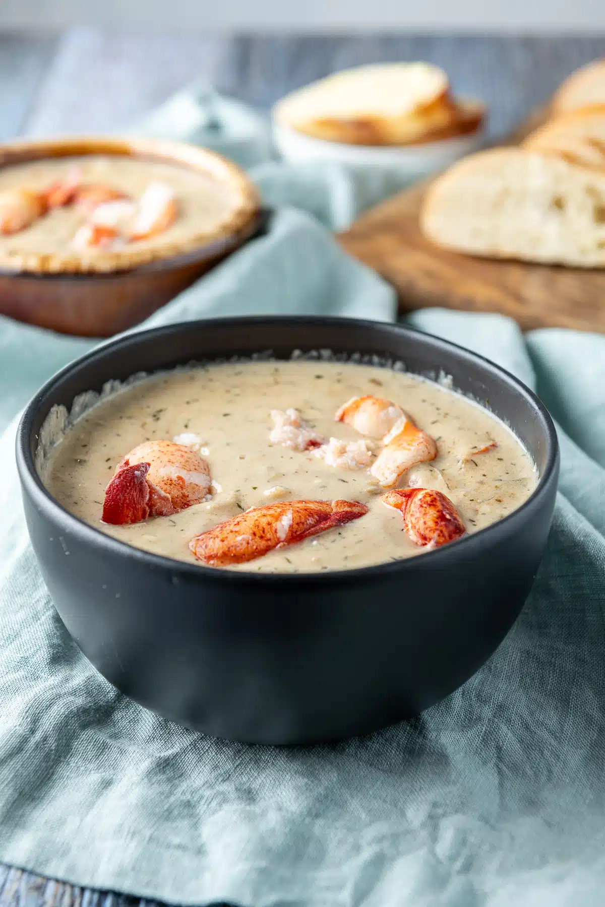 A black bowl filled with the creamy bisque with lobster on top and a crock in the back with bread