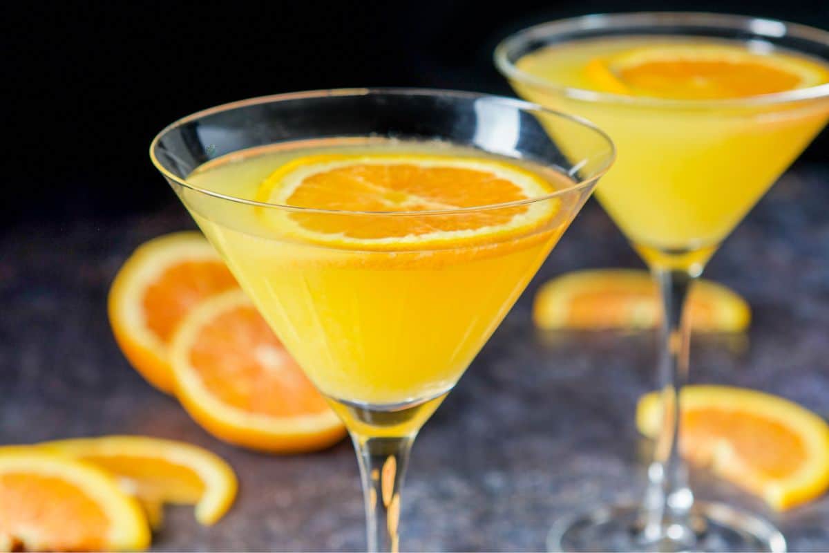 Two martini glasses filled with the orange martini with orange slices in the back