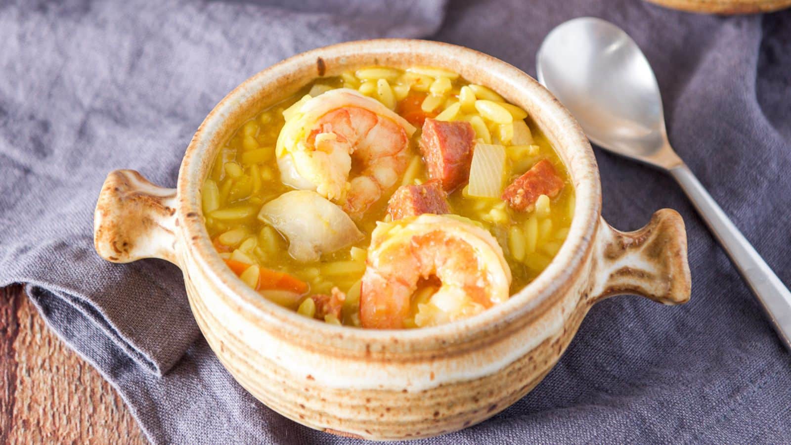 A grey napkin with a crock of soup on it. there are shrimp on top with chorizo on top