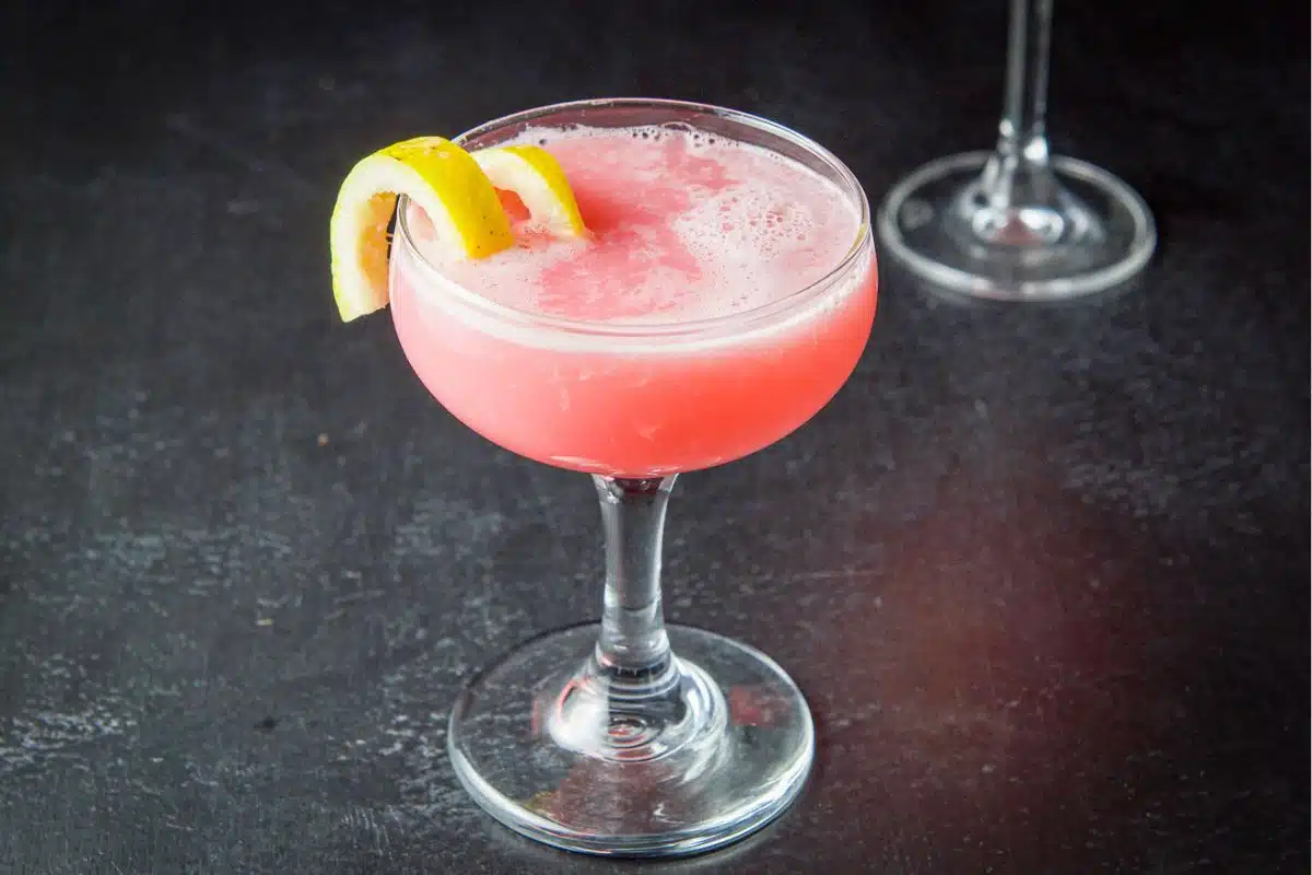 a coupe glass filled with a pink cocktail and a lemon twist on the side of the glass