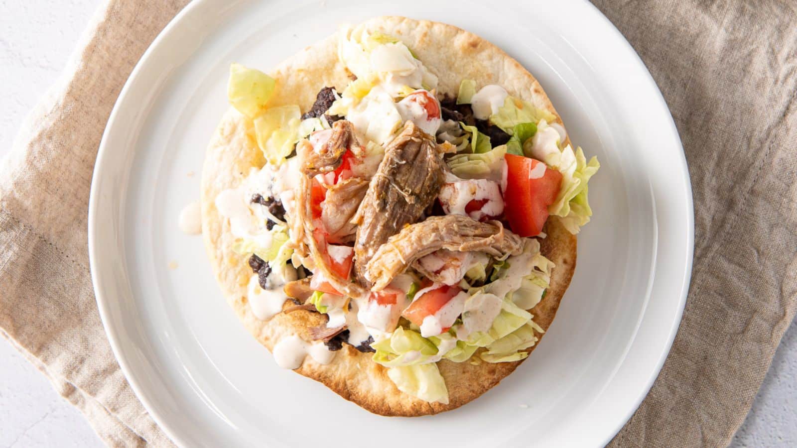 Overhead view of a crispy tortilla covered with lettuce, tomatoes, beans, a sauce and pulled pork