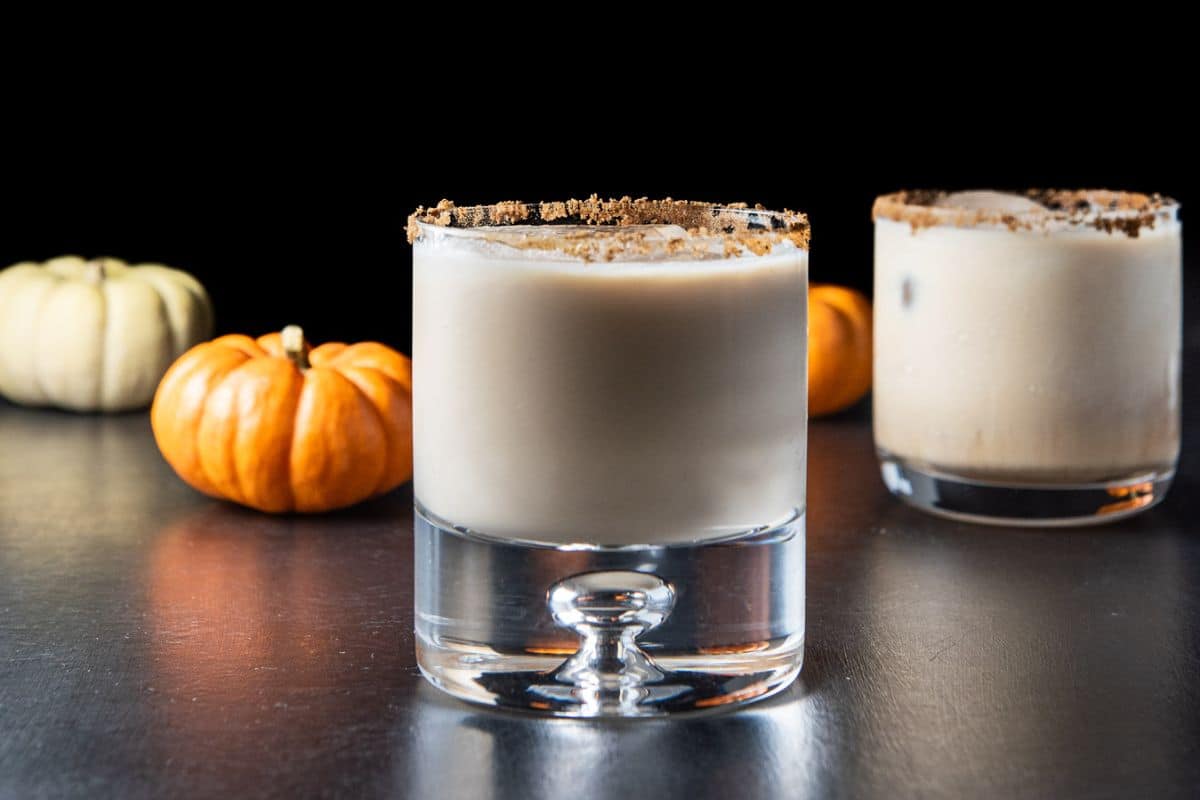 two glasses filled with a creamy cocktail with small pumpkins in the back