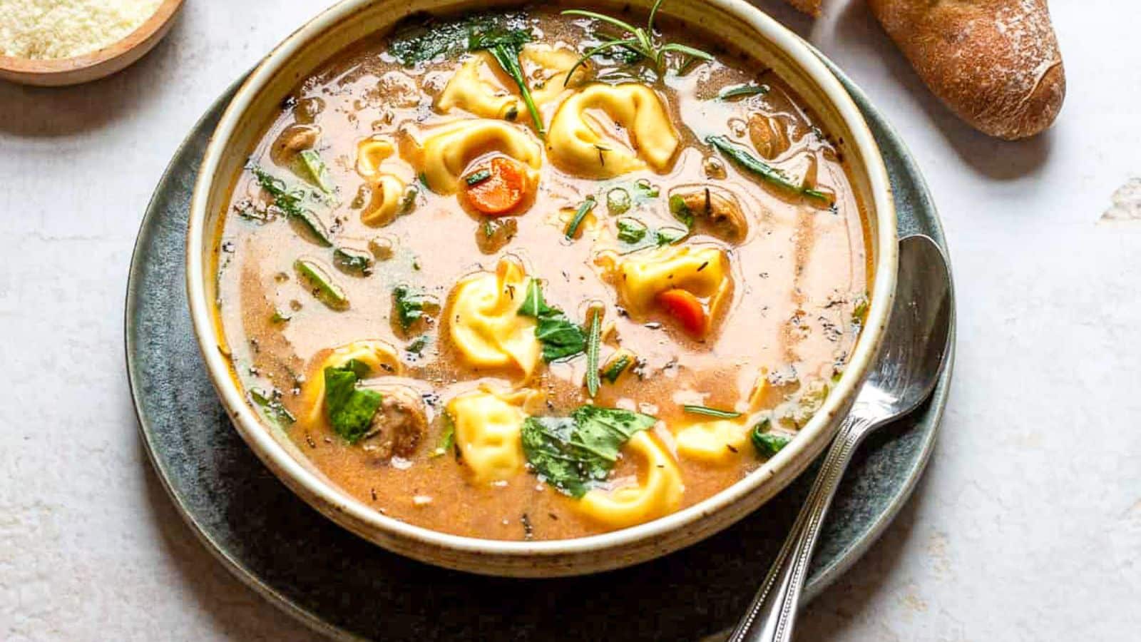 A brown bowl filled with tortellini soup on a plate with bread, and cheese