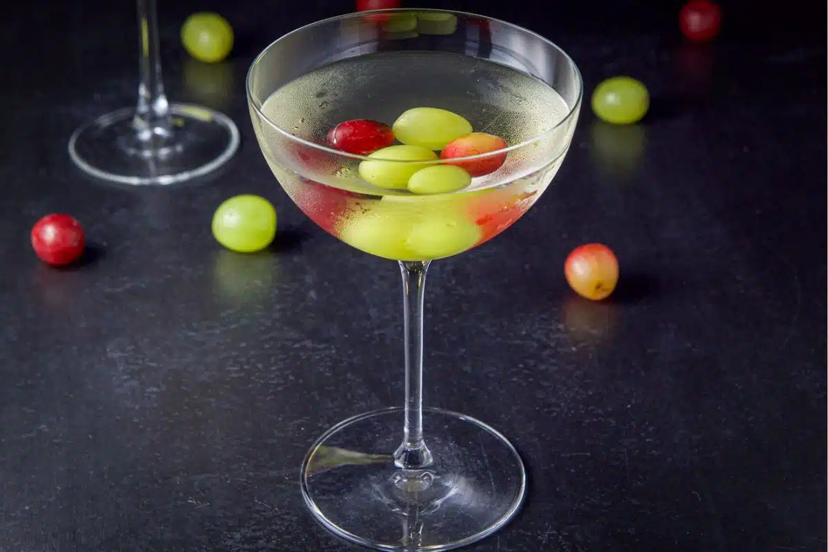 A bowl glass filled with a clear cocktail with grapes in the drink and on the table