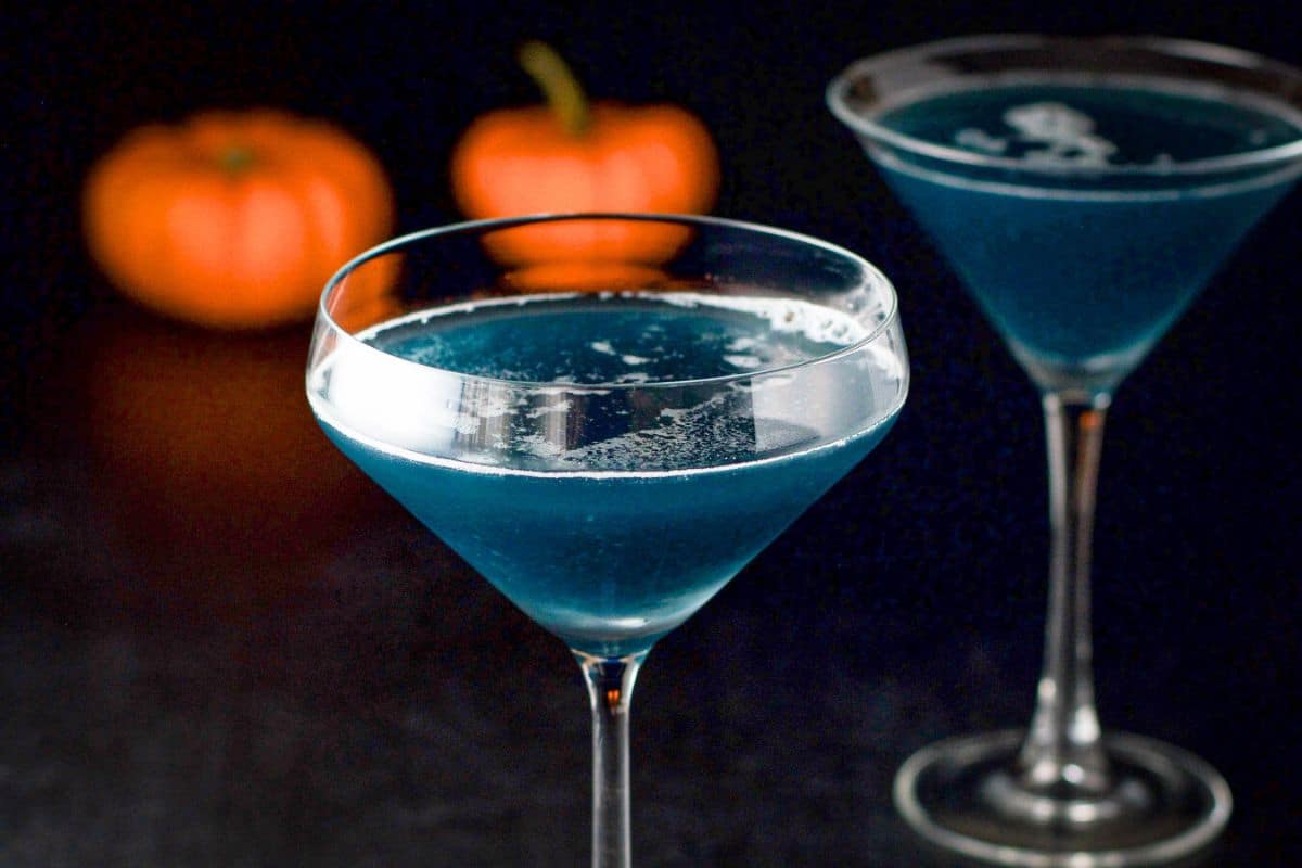 Two martini glasses with a blue cocktail in it; there are also small pumpkins in the back