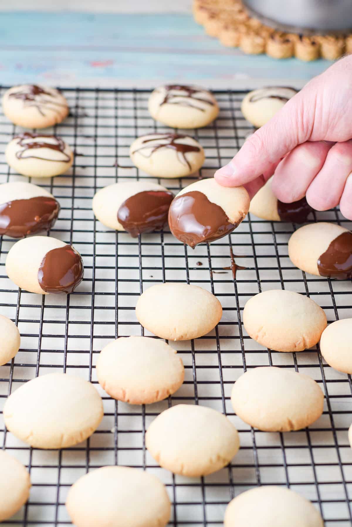 A hand holding a chocolate dipped cookie over a wire rack with a bunch of cookies on it.