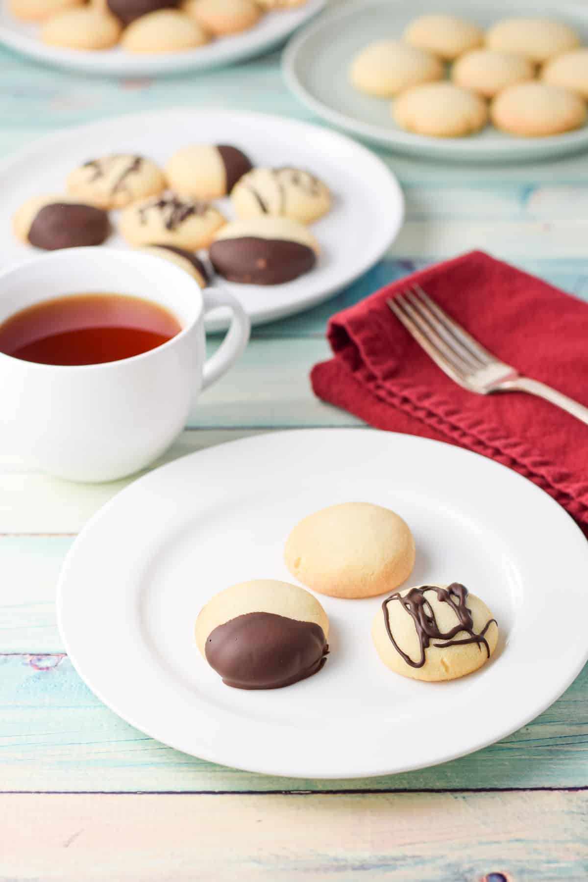 A few plates with shortbread cookies on it, both dipped and plain along with a cup of tea, napkin and fork