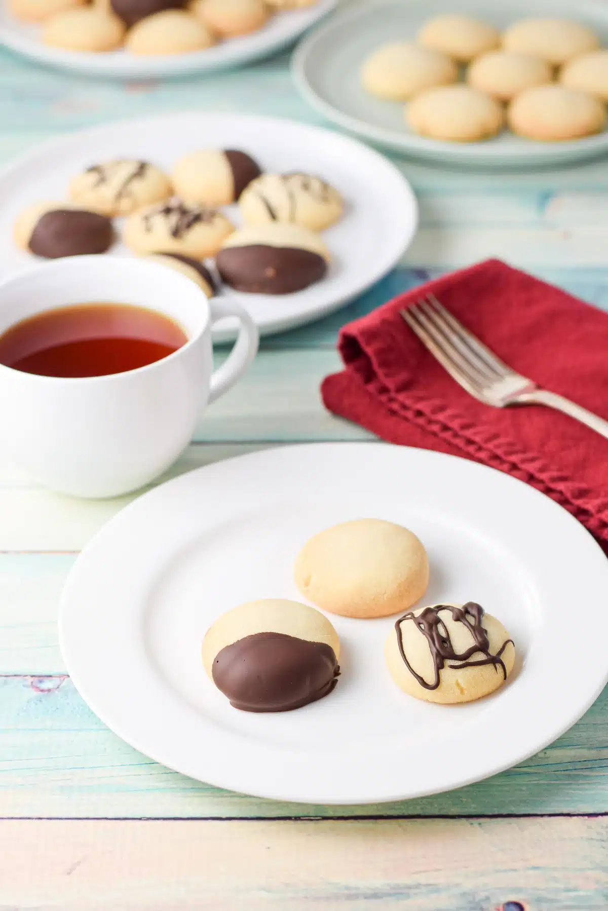 A few plates with shortbread cookies on it, both dipped and plain along with a cup of tea, napkin and fork