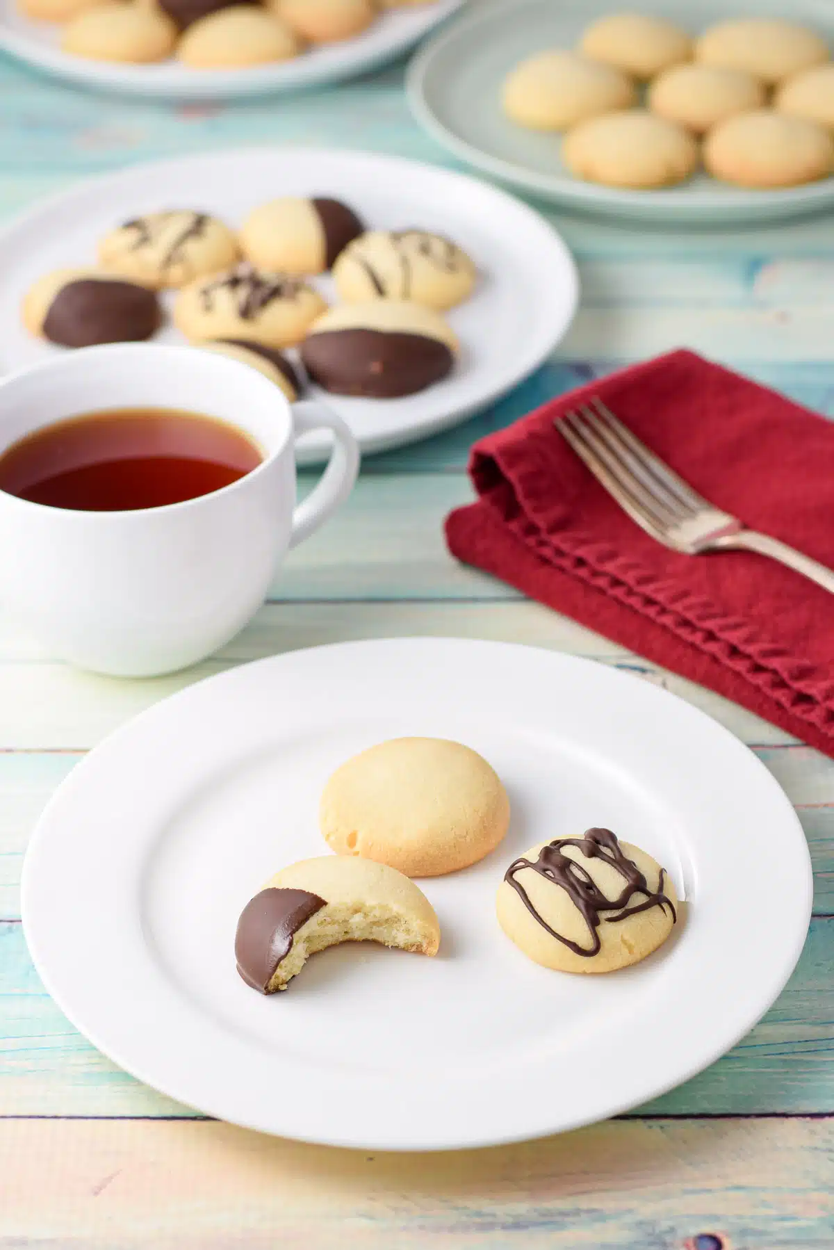 A white plate with 3 cookies on it with a bite out of one of them. There are other plates of cookies in back with a cup of tea