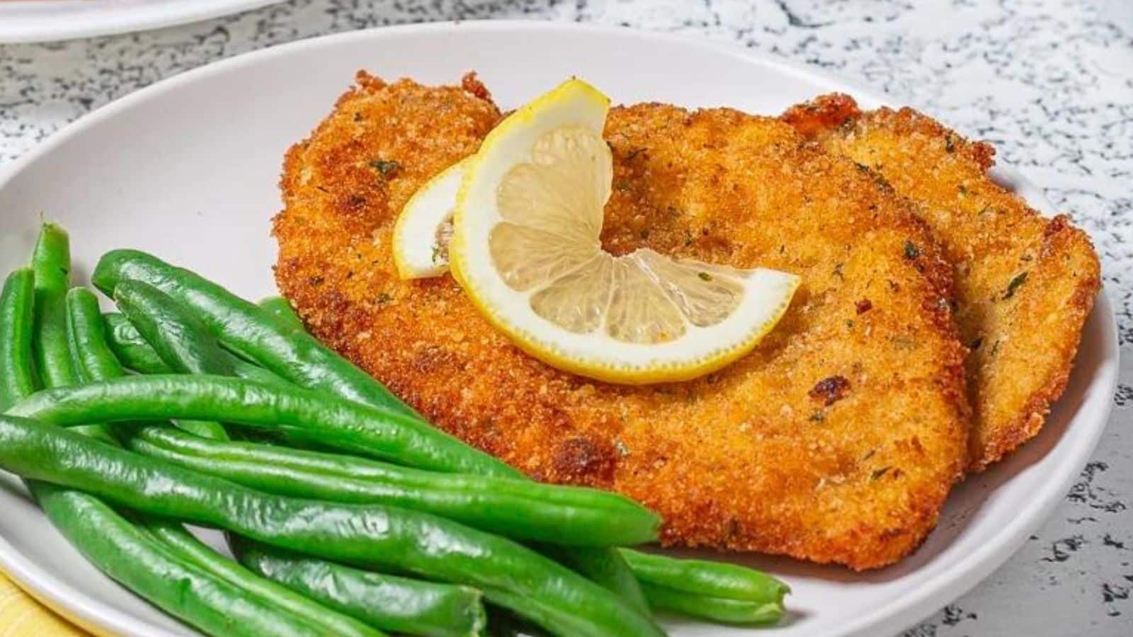A white plate with the cutlets on it with green beans and a slice of lemon