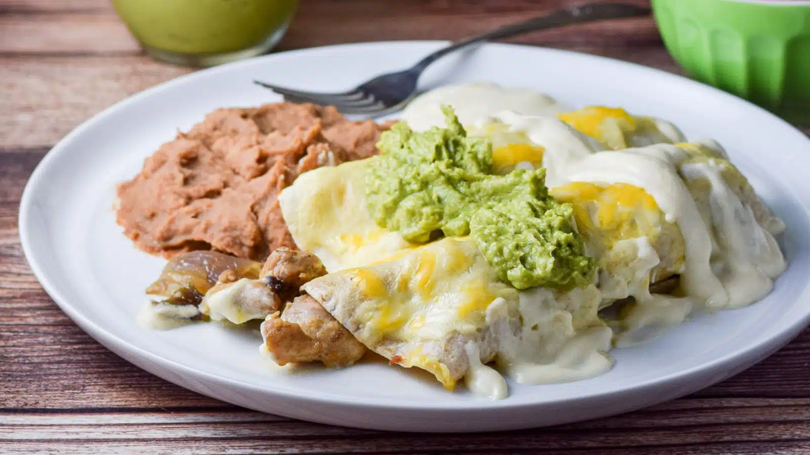 A white plate with two enchiladas with chicken sticking out of it with sauce and guacamole - along with beans on the plate