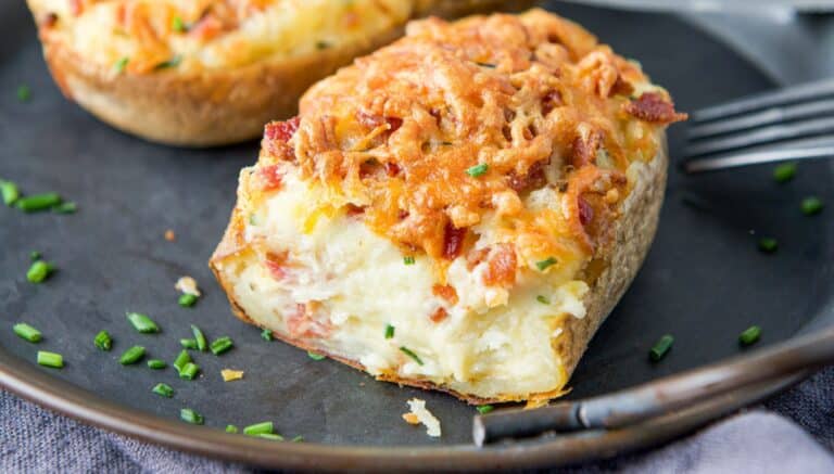A metal pan with two halves of baked potatoes with cheese and bacon on top. There are also chives on the tray