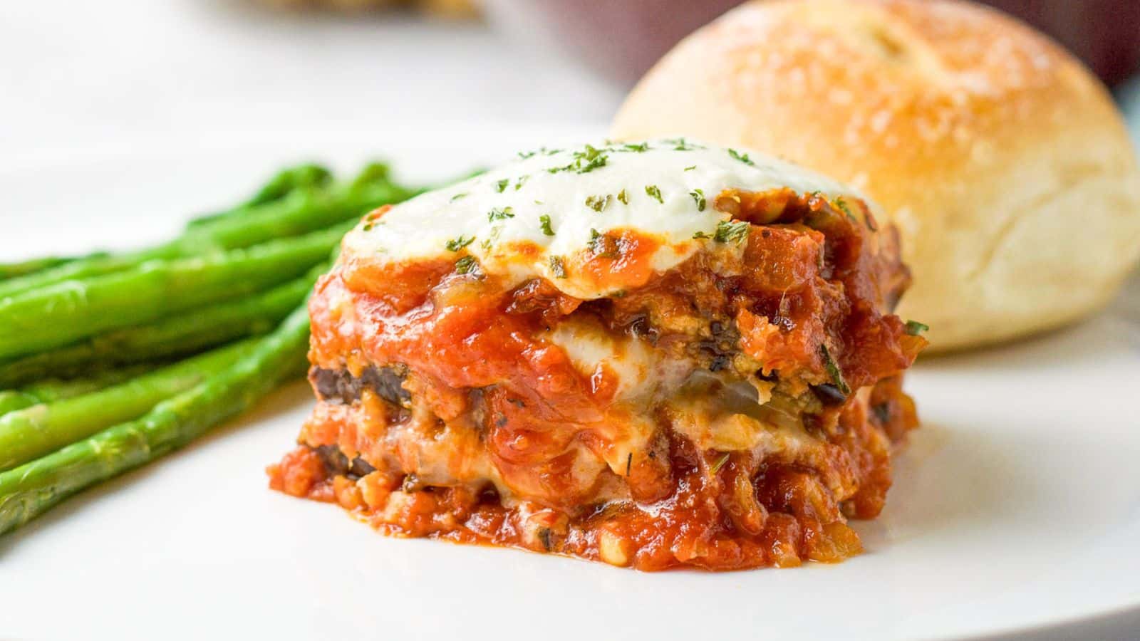 a close view of eggplant parmesan on a plate with asparagus and a roll