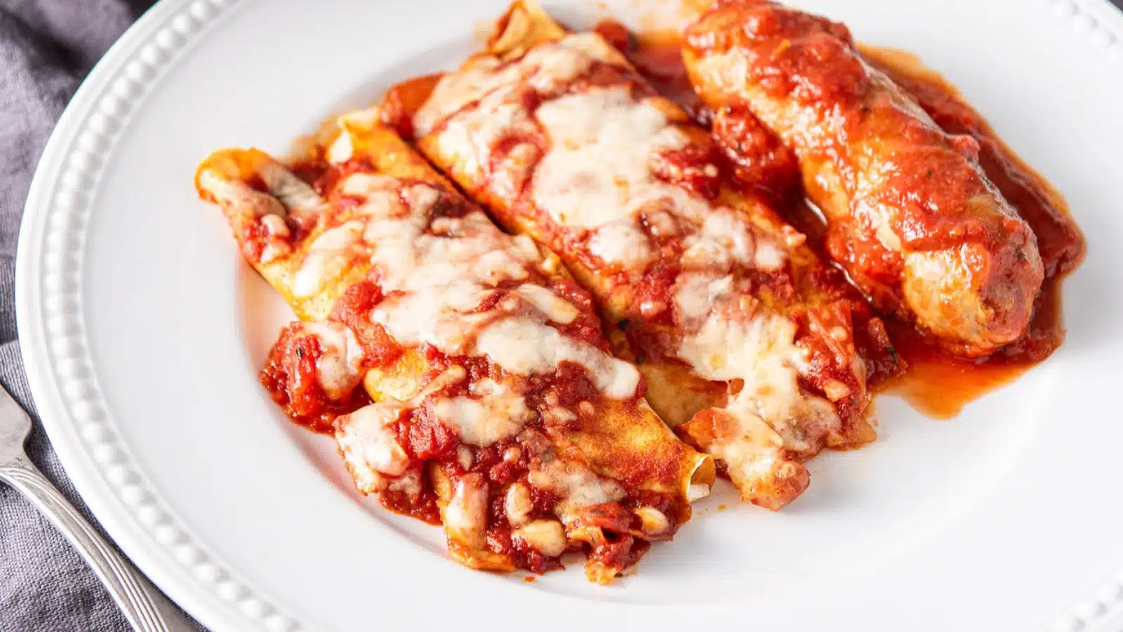 A beaded plate with two manicotti and a sausage on it with red sauce over it and melted cheese