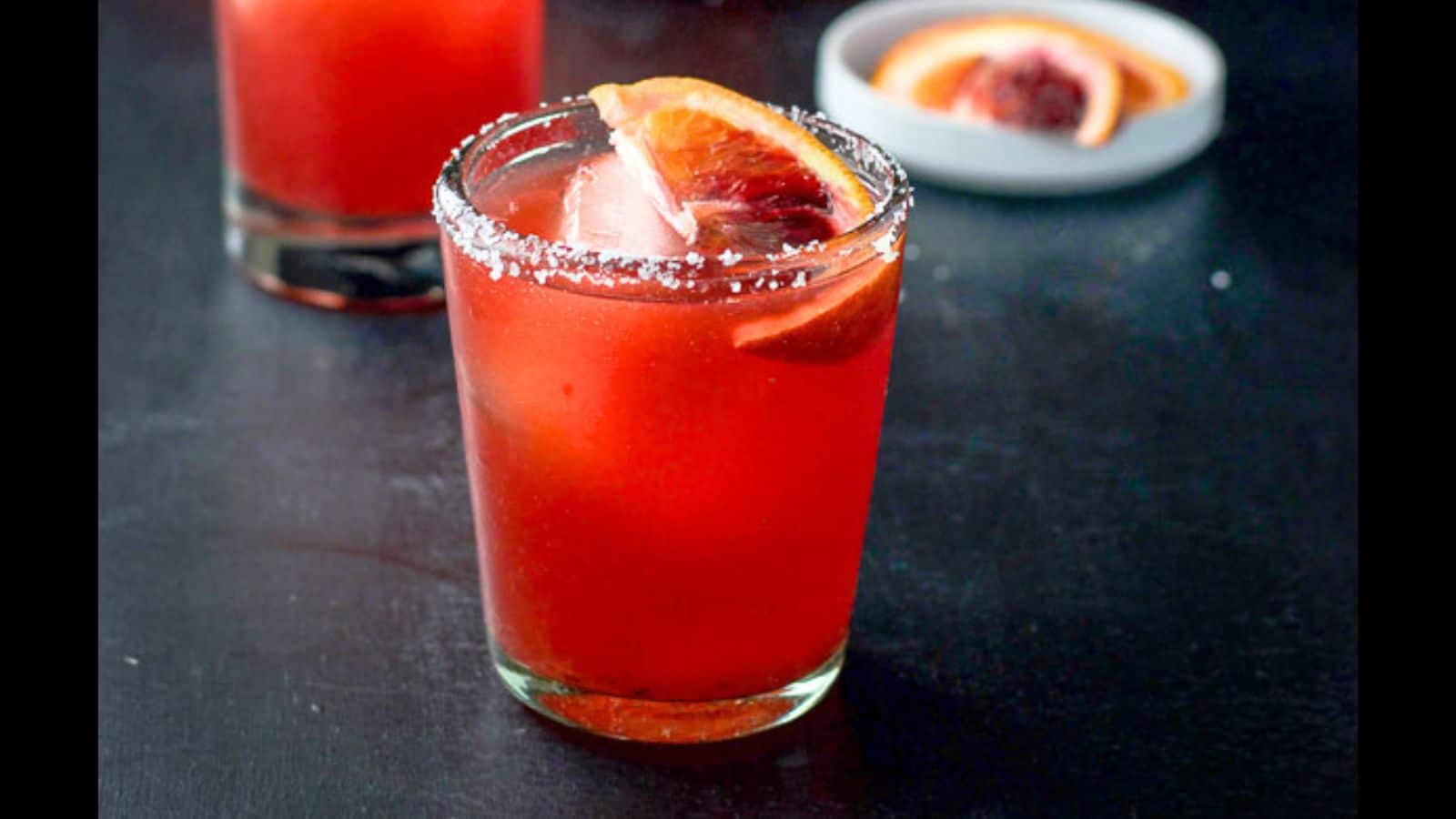 A glass with a salted rim with the blood orange drink in it along with a slice of orange in the drink and behind in a little white plate