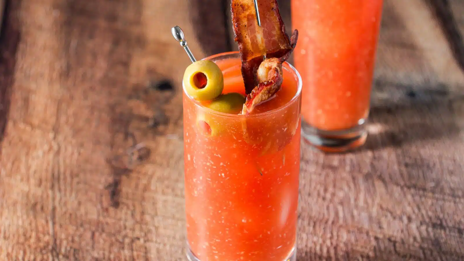 Two tall glasses with the bloody mary cocktail in it with olives and a piece of bacon