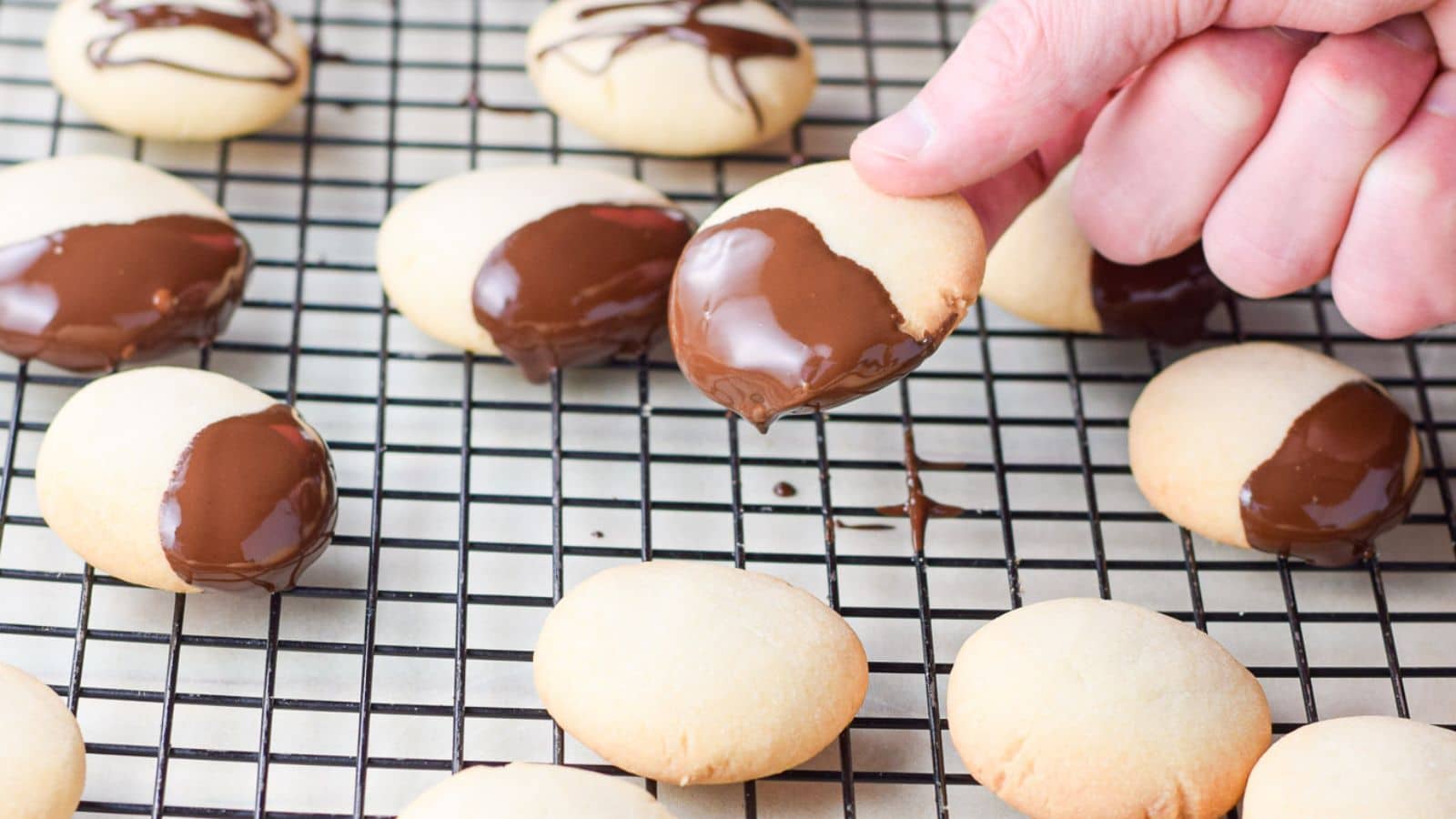 A male hand holding a cookie dipped in dark chocolate over a wire rack with other cookies on it