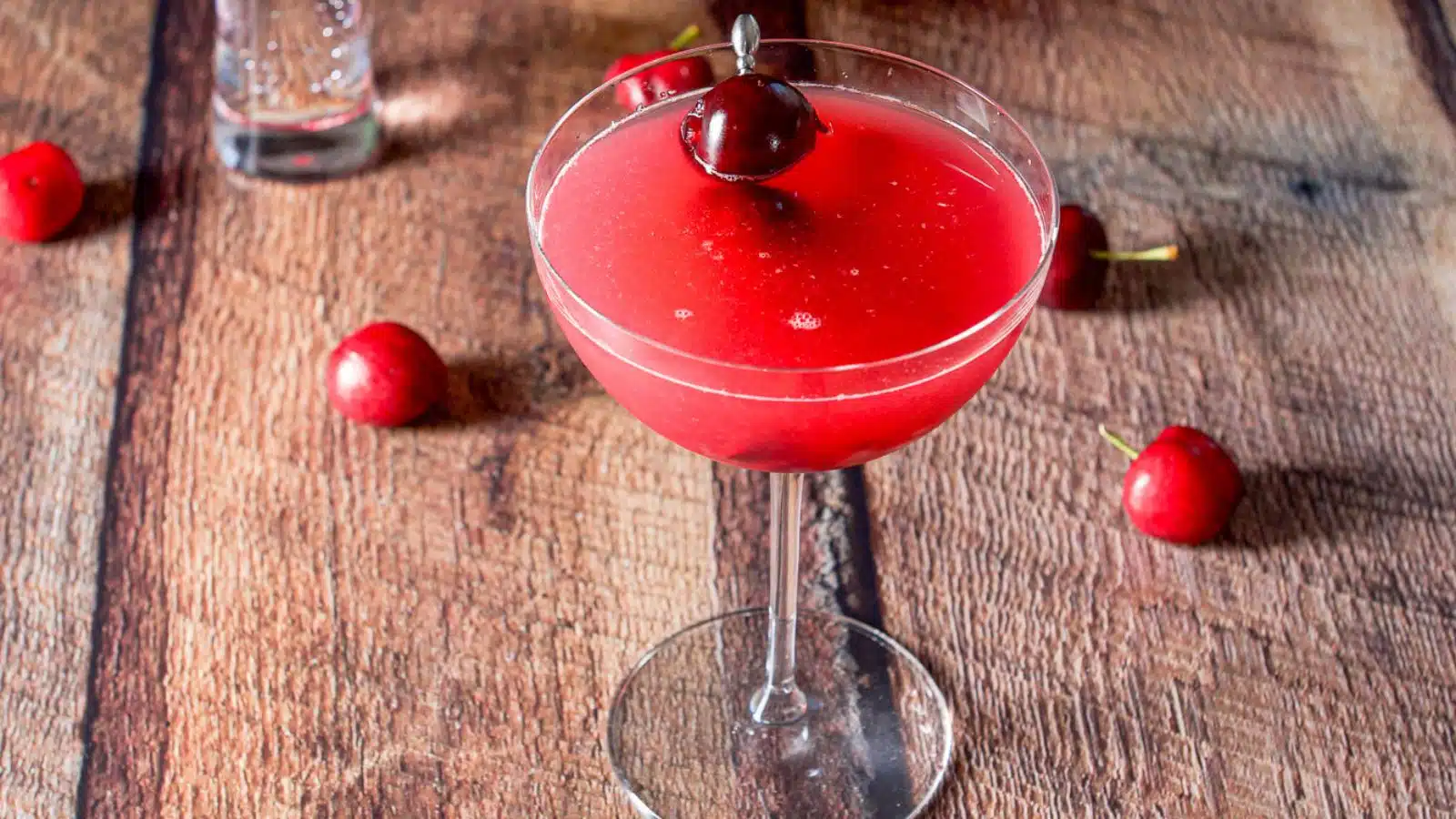 A bowl martini glass filled with a cherry cosmo with cherries on the table and as garnish