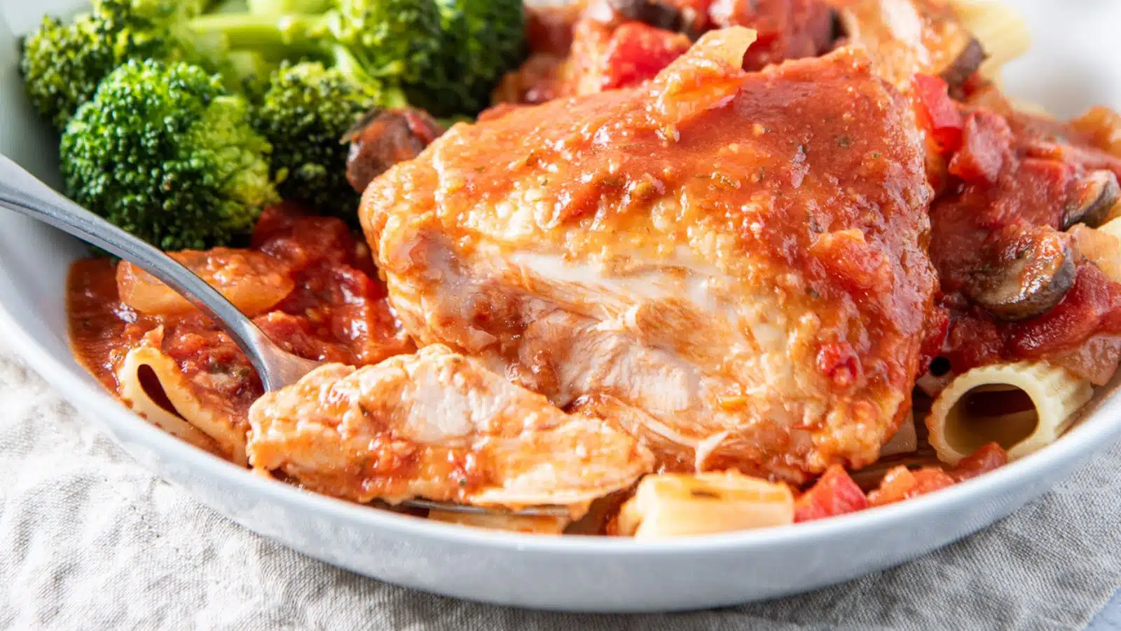 A white dish with a piece of chicken in red sauce on pasta with broccoli. There is a slice of chicken out of the thigh