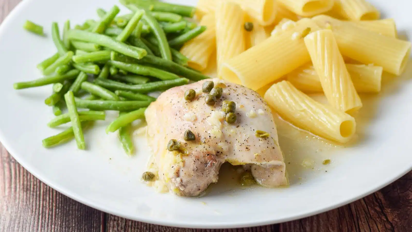 A white plate with a piece of chicken with capers and garlic with green beans and pasta and sauce