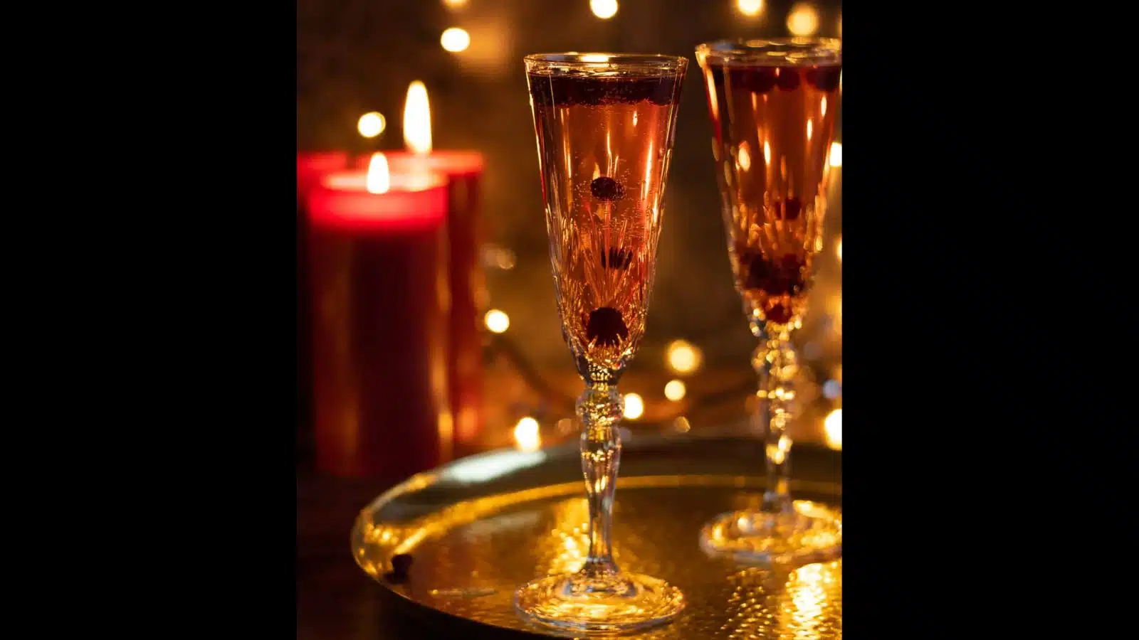 Two champagne flutes on a metal tray with champagne and cranberries. There are candles to the side